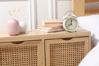 Yatağın yanında çalar saati, kitapları ve dekorasyonu olan bir komodin.
