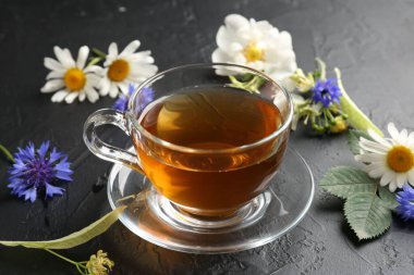Cam bardakta Aromatik bitki çayı, farklı çiçekler ve siyah masada yeşil yapraklar, yakın plan.