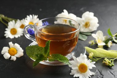 Cam fincanda aromatik bitki çayı, farklı çiçekler ve siyah masada nane şekeri, yakın plan.