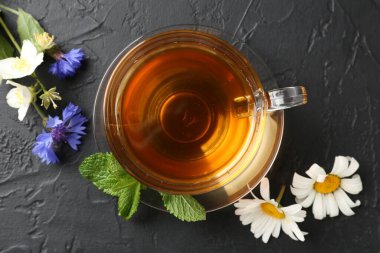 Cam fincanda aromatik bitki çayı, farklı çiçekler ve siyah masada nane şekeri.
