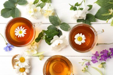 Cam fincanlarda aromatik bitki çayı, farklı çiçekler ve beyaz ahşap masada yeşil yapraklar.