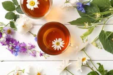 Cam fincanlarda aromatik bitki çayı, farklı çiçekler ve beyaz ahşap masada yeşil yapraklar.