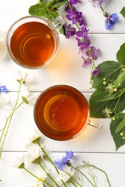 Cam fincanlarda aromatik bitki çayı, farklı çiçekler ve beyaz ahşap masada yeşil yapraklar.