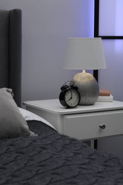 İçinde lamba, çalar saat ve yatağın yanında kitaplar olan komodin.