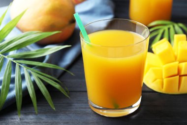 Bardakta lezzetli mango suyu, taze meyve ve ahşap masada yeşil yapraklar, yakın plan.