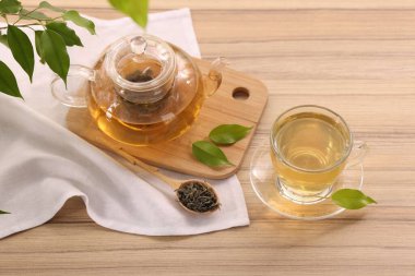 Cam fincanda lezzetli yeşil çay, çaydanlık, demlik ve yapraklar ahşap masada, manzaranın üstünde.