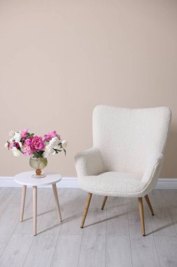 Bej duvarın yanında vazo dolusu çiçekli rahat bir koltuk ve masa.