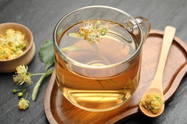 Koyu dokulu masa üzerinde tatlı ıhlamur çayı, kaşık, yaprak ve çiçekler, yakın plan.