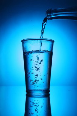 Mavi arkaplanda şişeden cama su dökmek. Arka ışıklandırma