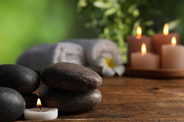 Spa composition with stones and burning candle on wooden table against blurred green background, closeup
