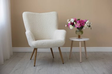 Bej duvarın yanında vazo dolusu çiçekli rahat bir koltuk ve masa.