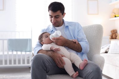 Baba küçük bebeğini evdeki koltukta şişeyle besliyor.