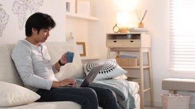 Yakışıklı Asyalı adam evdeki kanepede içip dizüstü bilgisayarla çalışıyor, kamera sola hareket ediyor.