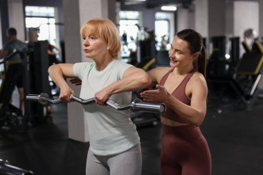 Kıdemli kadın spor salonunda profesyonel antrenör tarafından yönetilen EZ kıvrım çubuğuyla egzersiz yapıyor.