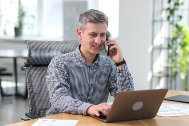 Gülümseyen işadamı akıllı telefondan konuşuyor ve ofiste dizüstü bilgisayarla çalışıyor.