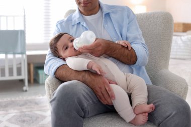 Babası küçük bebeğini evdeki koltukta biberonla besliyor.