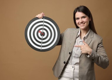Hedef alınıyor. Gülümseyen iş kadını bej arka planda dart tahtasını ve oku işaret ediyor.