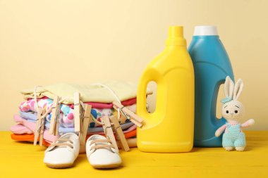 Kumaş yumuşatıcıları, bebek kıyafetleri, mandallar ve bej arka planda sarı ahşap masada oyuncaklar.