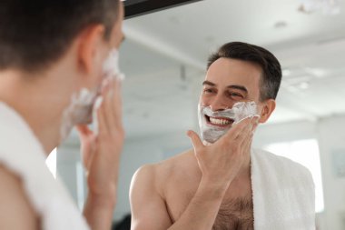 Gülümseyen adam evde aynanın yanında tıraş köpüğü sürüyor. Düşük açı.