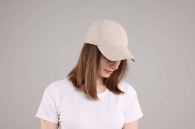 Açık gri arka planda bej renkli beysbol şapkası takan genç bir kadın.