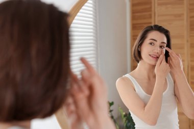 Kadın içeride aynanın yanına lens takıyor.