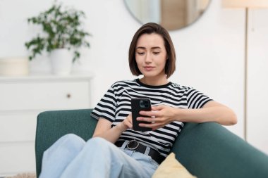 Evdeki koltukta modern akıllı telefon kullanan bir kadın.