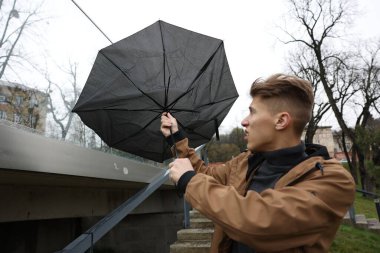 Kırık şemsiyeli bir adam açık havada yağmur altında rüzgarla savaşıyor.
