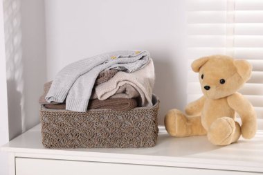 Wool sweaters in basket and teddy bear on white nightstand indoors