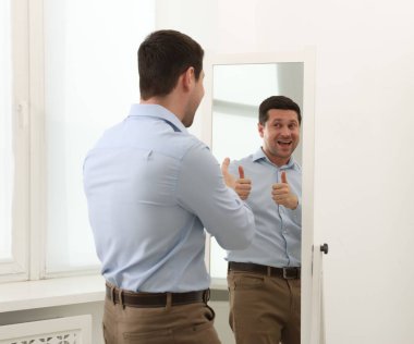 Kapalı alanda aynanın yanında baş parmağını gösteren yakışıklı adam.