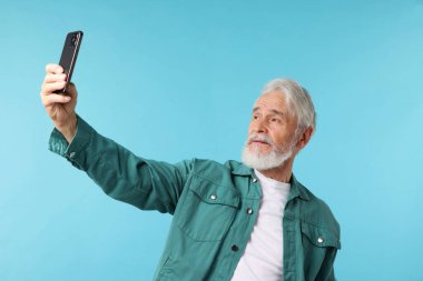 Açık mavi arka planda selfie çeken yaşlı bir adam