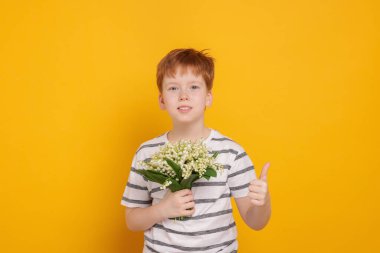 Elinde bir buket güzel çiçekli çocuk sarı arka planda baş parmağını gösteriyor.