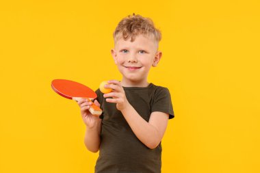 Sarı arka planda pinpon raketi ve topu olan küçük çocuk.