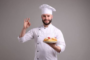 Üniformalı mutlu pastacı elinde çilekli lezzetli profiteroller tutuyor ve açık gri arka planda güzel bir jest yapıyor.