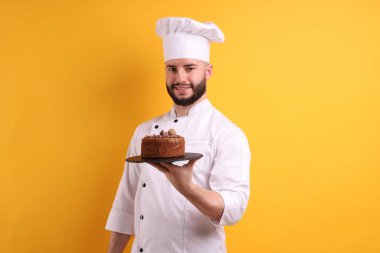 Üniformalı mutlu pastacı turuncu arka planda çikolatalı pasta tutuyor.
