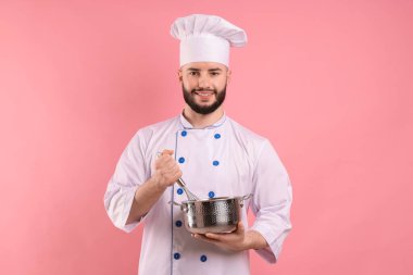 Üniformalı mutlu pastacı elinde çırpıcı ve pembe arka planda pişirme kabı tutuyor.