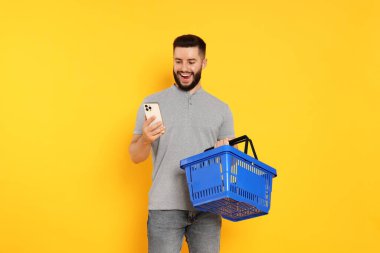 Boş alışveriş sepeti ve turuncu arka planda akıllı telefonu olan bir adam.