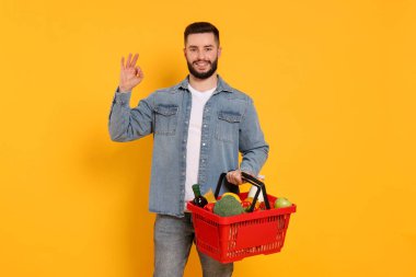 Elinde bir sepet dolusu ürün olan adam turuncu arka planda iyi bir jest gösteriyor.