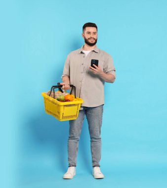 Ürünlerle dolu alışveriş sepeti ve açık mavi arka planda akıllı telefonu olan bir adam.