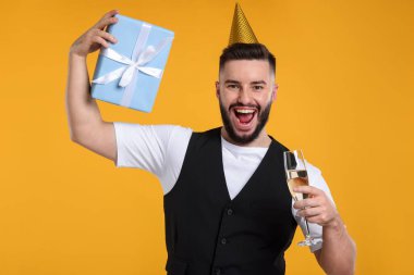 Doğum günün kutlu olsun. Parti şapkası, bir bardak şampanya ve turuncu arka planda hediye kutusu olan yakışıklı bir adam.