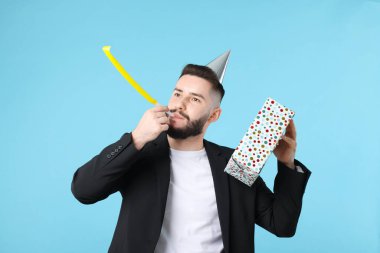Doğum günün kutlu olsun. Parti şapkası, üflemeli ve açık mavi arka planda hediye kutusu olan yakışıklı bir adam.