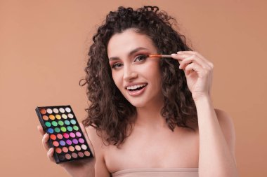 Göz farı paleti ve fırçası olan gülümseyen kadın bej arka planda makyaj yapıyor.