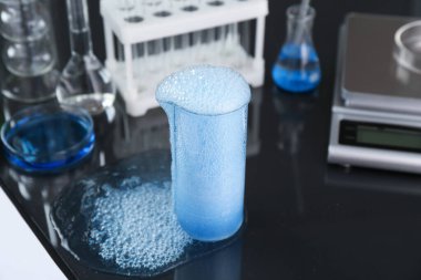 Mavi numuneli deney şişesi, diğer laboratuvar camları ve ölçekli siyah ayna masası, yakın plan.