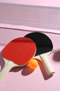 Masa tenisi raketleri, top ve net pembe arka planda, görünümün üstünde.