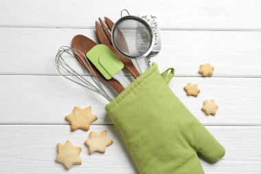 Fırın eldiveninde farklı mutfak aletleri ve beyaz ahşap masada yıldız şeklinde kurabiyeler.