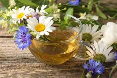 Cam bardakta Aromatik bitki çayı, tahta masada çiçekler ve yeşil yapraklar, yakın plan.