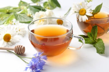 Cam fincanlarda aromatik bitki çayı, farklı çiçekler ve beyaz arkaplanda nane şekeri, yakın plan.