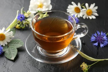 Cam bardakta Aromatik bitki çayı, farklı çiçekler ve siyah masada yeşil yapraklar, yakın plan.