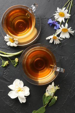 Cam fincanlarda aromatik bitki çayı ve siyah masada farklı çiçekler.