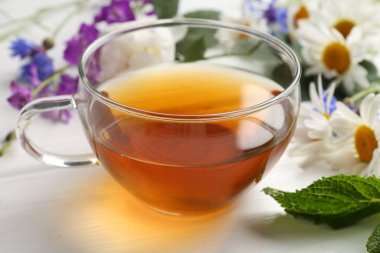 Cam bardakta Aromatik bitki çayı, farklı çiçekler ve beyaz ahşap masada nane şekeri, yakın plan.