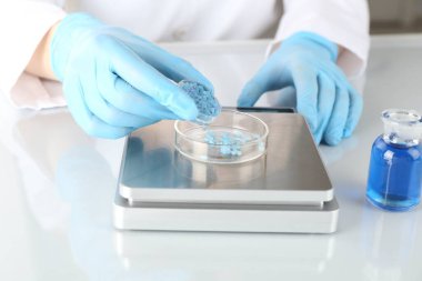 Bilim adamı beyaz ayna masasındaki terazide numune tartıyor, yakın plan.